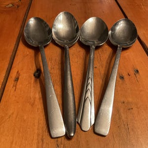 May include: Four stainless steel spoons with oval bowls and varying handle designs. The spoons are arranged on a wooden surface, showcasing their polished metal finish. The handles have different shapes and patterns.