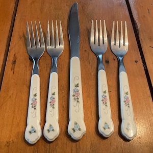 May include: A set of vintage silverware, including four forks and a knife. The handles are off-white with a floral design in pink and blue. The silverware is arranged on a wooden surface.