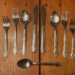 May include: A collection of vintage silverware, including forks, spoons, and knives, arranged on a wooden surface. The silverware has ornate, floral-like detailing on the handles. The spoons and forks have tines and bowls, while the knives have blades.