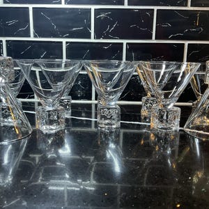 May include: A collection of six clear glass martini glasses. Each glass features a triangular bowl and rests on a decorative, square base. The glasses are arranged in a row, reflecting light and creating a sophisticated appearance.