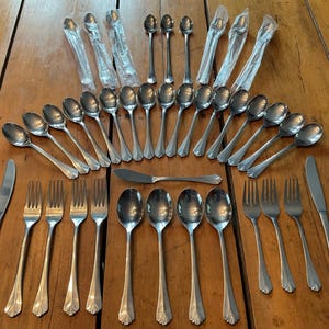 May include: A collection of stainless steel silverware, including spoons, forks, and knives, arranged on a wooden surface. Some spoons are still in their original plastic packaging. The silverware has a classic design.