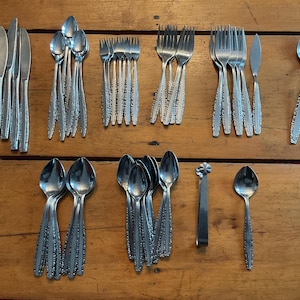 May include: A selection of stainless steel cutlery, including knives, forks, and spoons, arranged on a wooden surface. The cutlery features a decorative pattern on the handles. A serving utensil is also included.
