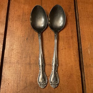 May include: Two silver spoons with ornate handles are displayed against a wooden background. The spoons have a classic design, with a rounded bowl and a detailed floral pattern on the handle. The spoons are likely for serving or eating.