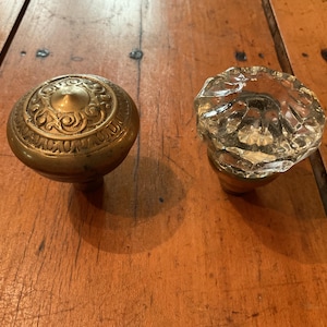 May include: Two door knobs are displayed side by side. One knob is crafted from brass with a circular, embossed pattern on its top surface. The second knob is made of clear, faceted glass with a multi-sided, reflective design and a metallic base.
