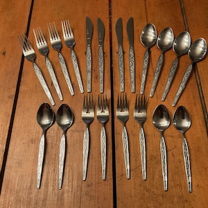May include: A collection of stainless steel silverware, including forks, knives, and spoons, arranged on a wooden surface. The silverware features a decorative floral pattern on the handles. The spoons have a rounded bowl shape.