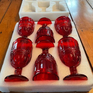 May include: Six ruby red wine glasses with ornate detailing are arranged in a white foam insert. The glasses have a classic design with a rounded bowl and a stemmed base. The glasses are likely made of glass.