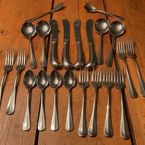 May include: A collection of vintage silverware, including spoons, forks, and knives, arranged on a wooden surface. The silverware has a shiny, metallic finish. The arrangement suggests a formal dining setting.