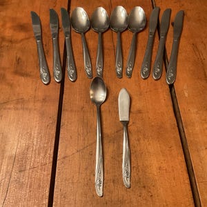 May include: A collection of vintage silverware, including spoons, knives, and a butter knife, arranged on a wooden surface. The silverware has a floral design on the handles. The spoons and knives are arranged in a fan shape.