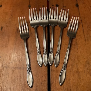 May include: Six silver forks with decorative floral detailing on the handles are arranged on a wooden surface. The forks have four tines each and are made of a shiny, reflective metal.