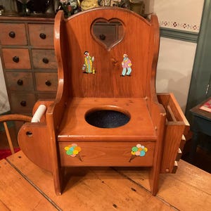 May include: A vintage wooden potty chair with a heart-shaped cutout in the backrest. The chair features colourful clown stickers and a small storage compartment. The wood has a warm, reddish-brown tone.