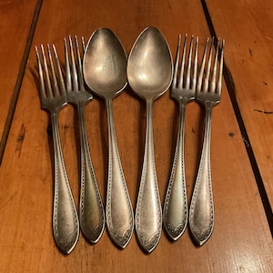 May include: Six pieces of antique silverware, including two spoons and four forks, are arranged on a wooden surface. The silverware has a silver finish with decorative detailing on the handles. The spoons and forks are placed in an alternating pattern.