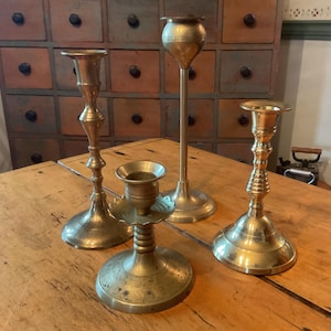 May include: Four antique brass candle holders of varying heights and designs sit on a wooden table. The candle holders have a warm, golden hue and show signs of age. The background includes a wooden cabinet with multiple drawers.