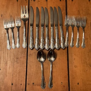 May include: A collection of vintage silverware arranged on a wooden surface. The set includes forks, knives, and spoons with ornate handles. The silverware is made of a shiny, silver-colored metal, reflecting the light. The arrangement suggests a formal dining setting.