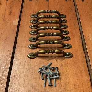 Peut inclure: Ensemble de sept poignées de tiroir en laiton antique avec vis assorties, disposées sur une surface en bois. Les poignées ont un design incurvé avec des détails décoratifs, et les vis sont argentées.