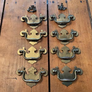 May include: Eight antique brass drawer pulls with matching screws are arranged on a wooden surface. The pulls have a curved handle design with decorative backplates. The brass has a warm, aged patina.