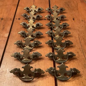 May include: A collection of twelve antique brass drawer pulls. Each pull features a decorative, ornate design with a central flourish and curved handles. The brass has a warm, aged patina, suggesting vintage craftsmanship.