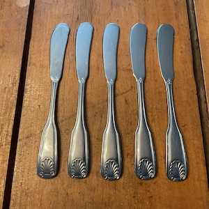 May include: Five silver butter knives with curved blades and decorative shell designs on the handles are arranged on a wooden surface. The knives are made of a shiny, metallic material.