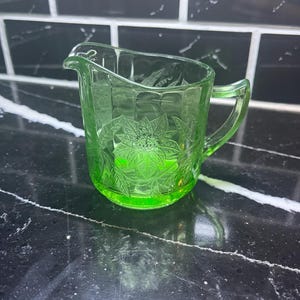 May include: A green glass creamer with a handle and a spout. The creamer has an embossed floral design on the front. The glass has a slight green tint, and the creamer is on a black surface.