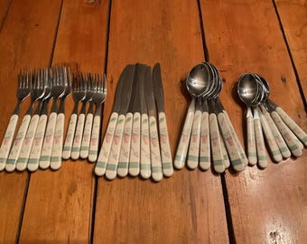May include: A collection of vintage silverware, including forks, knives, and spoons. The handles feature a cream-colored base with a floral and gardening tool design in pink, green, and brown. The silverware is arranged on a wooden surface.