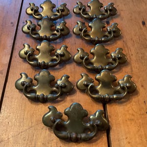 May include: A collection of nine matching brass drawer pulls arranged in rows. Each pull features a decorative backplate with a central fleur-de-lis shape and a curved, swinging handle attached to two points, showcasing a dark, aged metallic finish.