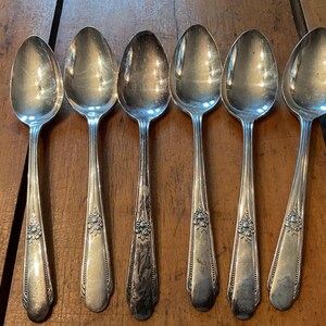 May include: Six silver spoons on a wooden surface. The spoons have oval bowls and floral patterns on the handles. The spoons reflect the surroundings. The spoons are arranged in a row.