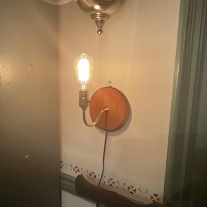 May include: A wall-mounted light fixture with a wooden backplate and a vintage-style Edison bulb. The fixture has a brass-colored arm and a brown electrical cord. The light is turned on, illuminating the surrounding wall.