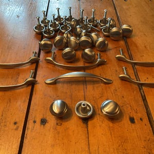 May include: A collection of brushed nickel cabinet hardware including multiple round knobs and several arched drawer pulls. The hardware features a smooth, metallic finish with a consistent silver tone, designed for attachment to furniture or cabinetry.