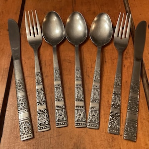 May include: A collection of six pieces of vintage silverware, comprising two forks, two spoons, and two knives. The silver cutlery features decorative floral and geometric designs on the handles. The set is displayed on a wooden surface.