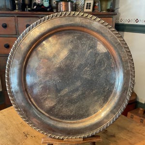 May include: A large, round silver serving tray with an ornate, embossed floral design in the center. The tray has a decorative, rope-like edge. The tray is displayed on a wooden surface.