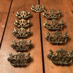 Peut inclure: Ensemble de poignées de tiroir antiques en laiton, disposées sur une surface en bois. Les poignées présentent un design orné et symétrique avec des poignées incurvées et des détails décoratifs. Le laiton a une patine chaude et vieillie.