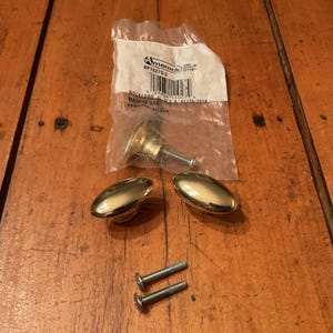 May include: Three oval-shaped brass cabinet knobs with a polished finish. One knob is inside a clear plastic Amerock brand packaging bag with a barcode and text. Two additional knobs are loose, accompanied by two silver metal mounting screws.