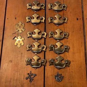 May include: A collection of ten brass drawer pulls with a decorative bail handle design, two matching brass keyhole escutcheons, and a small pile of metal mounting screws. The hardware features a polished metallic finish with ornate detailing.
