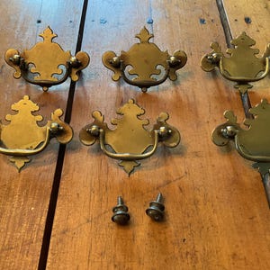 May include: Six antique brass drawer pulls with ornate designs and two matching screws are arranged on a wooden surface. The pulls have a curved handle and decorative backplate, showcasing a vintage aesthetic.