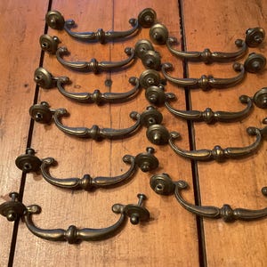 May include: A collection of ten antique-style brass drawer pulls arranged in two columns. Each pull features a curved handle with a central decorative detail and two circular mounting plates at the ends, showcasing a dark, aged metallic finish.