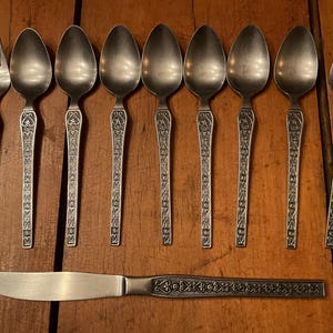 May include: A collection of vintage silverware, including spoons, forks, and a knife, arranged on a wooden surface. The silverware has ornate, detailed handles with a floral design. The spoons and forks have a rounded bowl and tines, respectively.