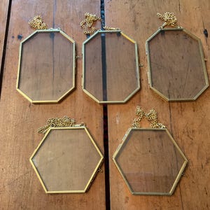 May include: Five vintage gold-coloured metal and glass picture frames with decorative chain hangers. The frames have geometric shapes, including octagons and hexagons. The glass is clear, set against a wooden surface.