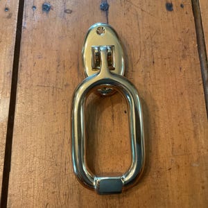 May include: A polished brass door knocker, featuring an oval backplate and a large, rounded rectangular handle. The knocker is mounted on a wooden door, highlighting its reflective surface and classic design. This door knocker is a decorative hardware element.