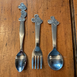 May include: Three stainless steel utensils: a spoon, a fork, and a spoon. Each utensil features a Mickey Mouse head or full figure design at the top of the handle. The utensils are set against a wooden surface.