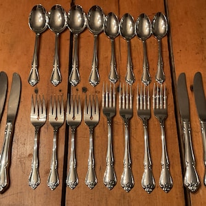 May include: A set of silver-coloured cutlery, including spoons, forks, and knives, arranged on a wooden surface. The silverware has ornate detailing on the handles. The set includes various sizes of spoons and forks, along with several knives.