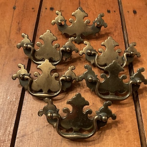May include: A collection of antique brass drawer pulls. The pulls have a decorative, ornate design with a curved handle and detailed backplate. The hardware is arranged on a wooden surface.