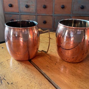 May include: Two copper-colored mugs with brass handles sit on a wooden surface. Each mug has a rounded shape and a shiny finish. The letter "P" is etched on the front of each mug. The mugs are likely for beverages.