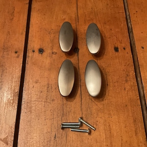 May include: Four oval-shaped cabinet pulls with a brushed metal finish, arranged in a square formation. Several silver screws are positioned below the pulls. The hardware is set against a warm, wooden surface.