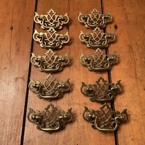 May include: Eight antique brass drawer pulls with ornate, symmetrical designs. Each pull features a central decorative element with curved handles and intricate detailing. The pulls are arranged on a wooden surface, showcasing their vintage aesthetic.