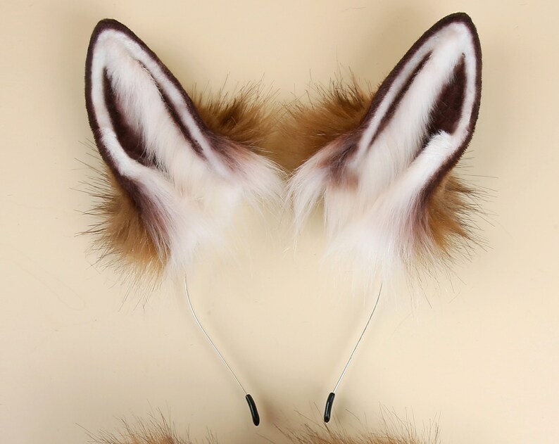 May include: A pair of brown and white faux fur fox ears with a silver headband. The ears are attached to the headband with black clips.
