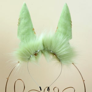 May include: A pair of green furry cat ears with gold accents on a headband. The ears are made of soft, plush fabric and have a realistic look.