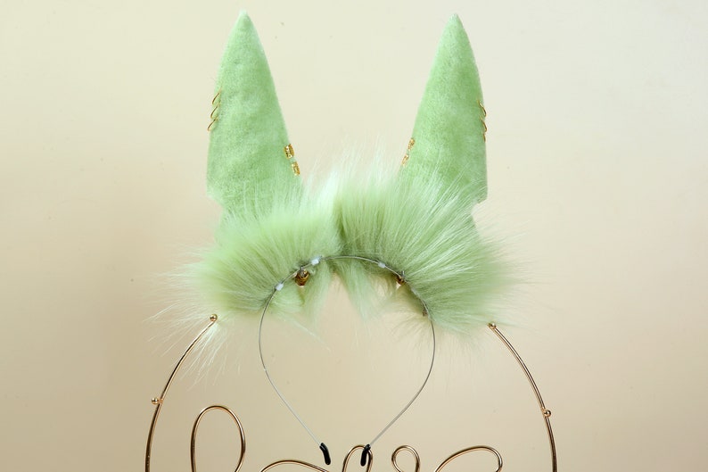 May include: A pair of green furry cat ears with gold accents on a headband. The ears are made of soft, plush fabric and have a realistic look.