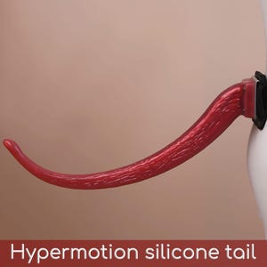 May include: A red silicone tail with a textured surface attached to a black adjustable strap. The tail has a curved, tapered shape and is mounted on a black base. The text "Hypermotion silicone tail" is printed at the bottom of the image.