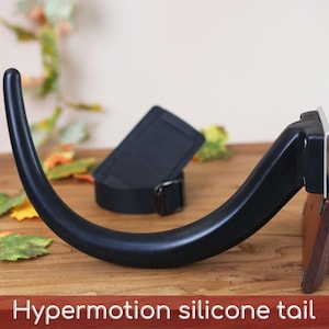 May include: A black silicone tail with a curved, tapered shape attached to a dark wood base with a metal bracket. A black fabric belt with a metal D-ring buckle is positioned behind the tail. The text "Hypermotion silicone tail" is visible.