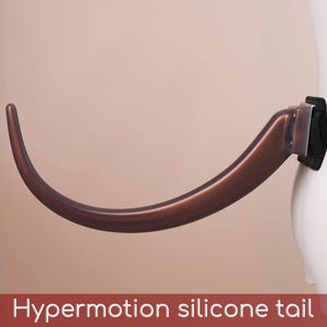 May include: A brown, curved silicone tail accessory attached to a black nylon belt. The tail features a smooth, tapered design and is connected to a rigid base mount. The text "Hypermotion silicone tail" is printed at the bottom of the image.