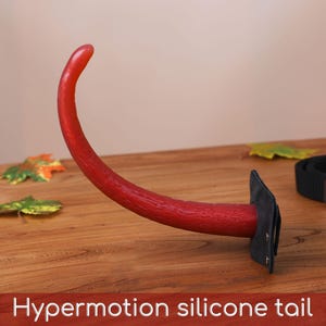 May include: A red, curved silicone tail with a textured surface, attached to a black rectangular base with metal rivets. The tail is positioned upright, and the text "Hypermotion silicone tail" is visible at the bottom of the image.